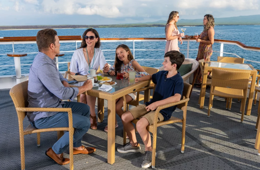 Family of four enjoying an al fresco lunch on the open deck of Yacht La Pinta with volcanic Galapagos coastline visible in the background.
