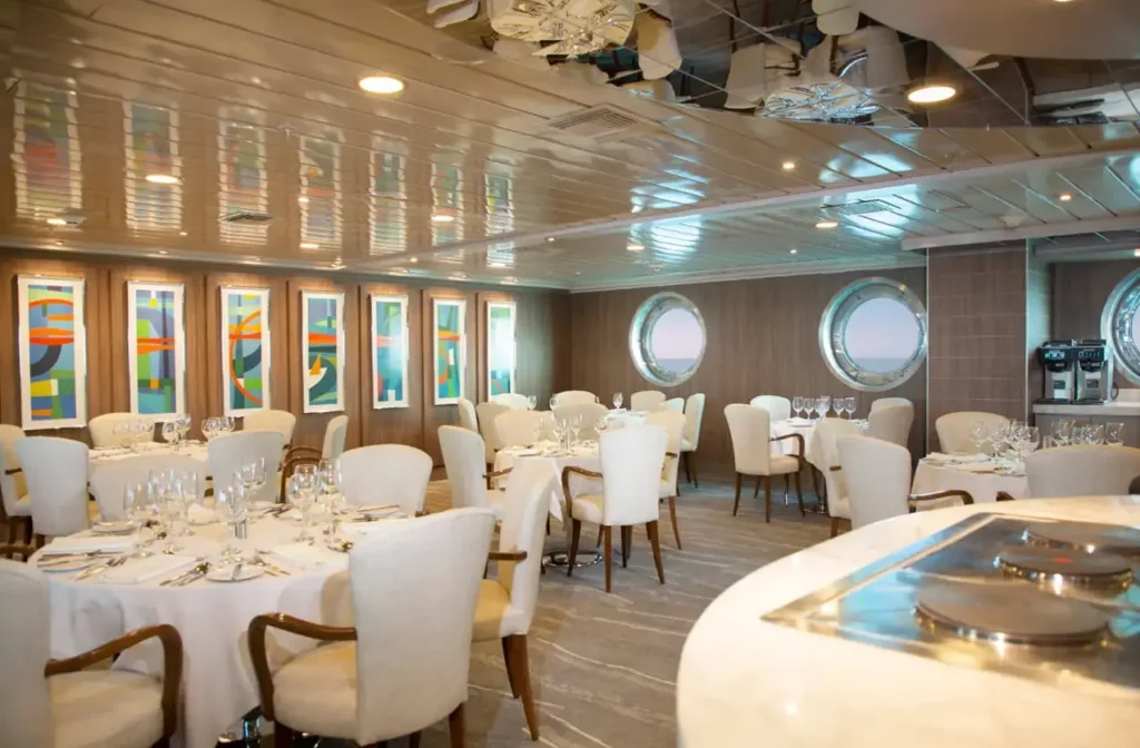 Elegant dining room aboard Yacht La Pinta with white upholstered chairs, formally set tables, colorful artwork and round porthole windows overlooking the ocean.
